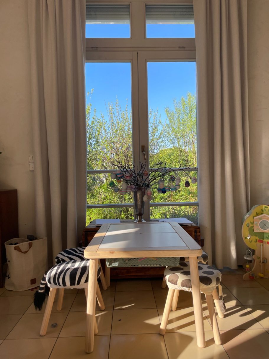 Ein heller Raum mit einem Tisch, zwei Stühlen und Blick auf grüne Bäume durch ein Fenster.
