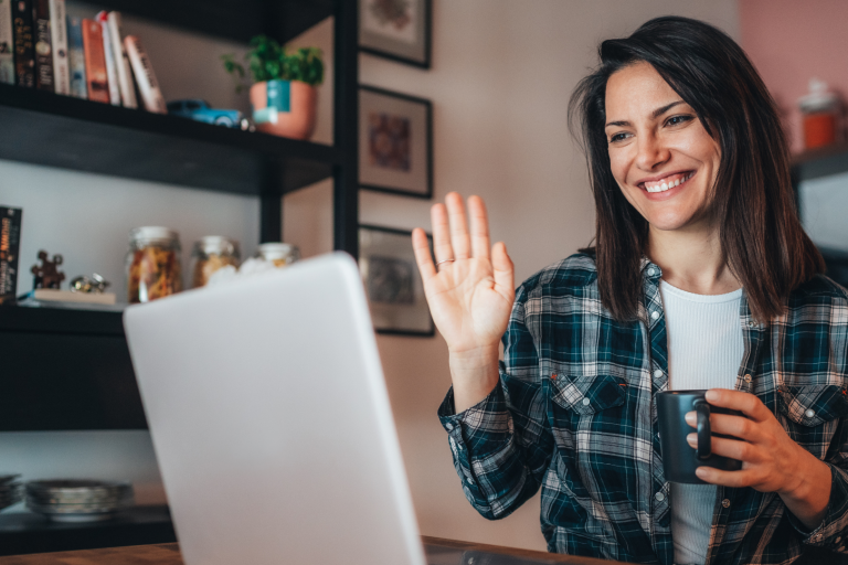 Frau mit Kaffeetasse winkt lächelnd einem Laptop zu.