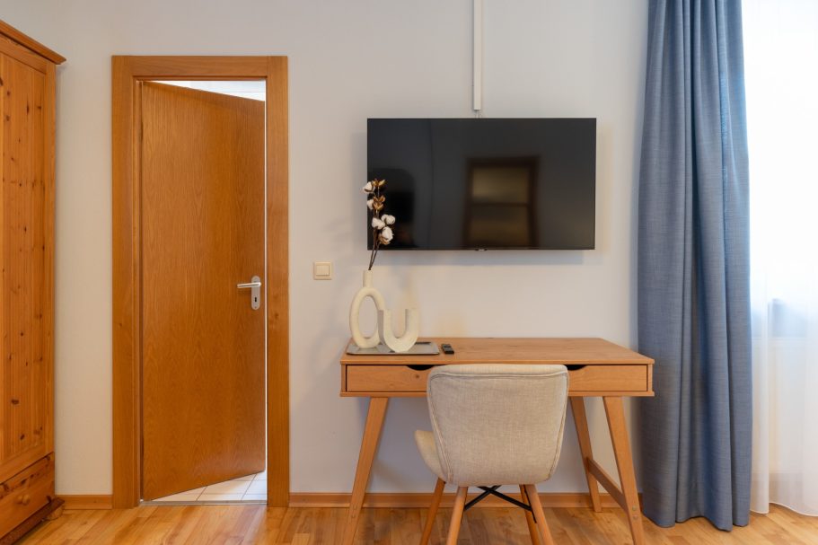 Heller Raum mit Schreibtisch, Stuhl und Wand-TV neben einer Holzinnentür.