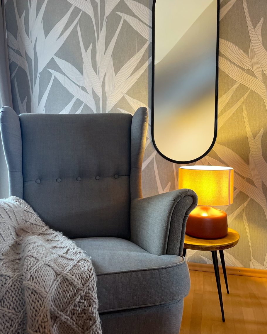 Grauer Sessel mit einer Decke, neben einer Lampe und einem ovalen Spiegel an der Wand.
