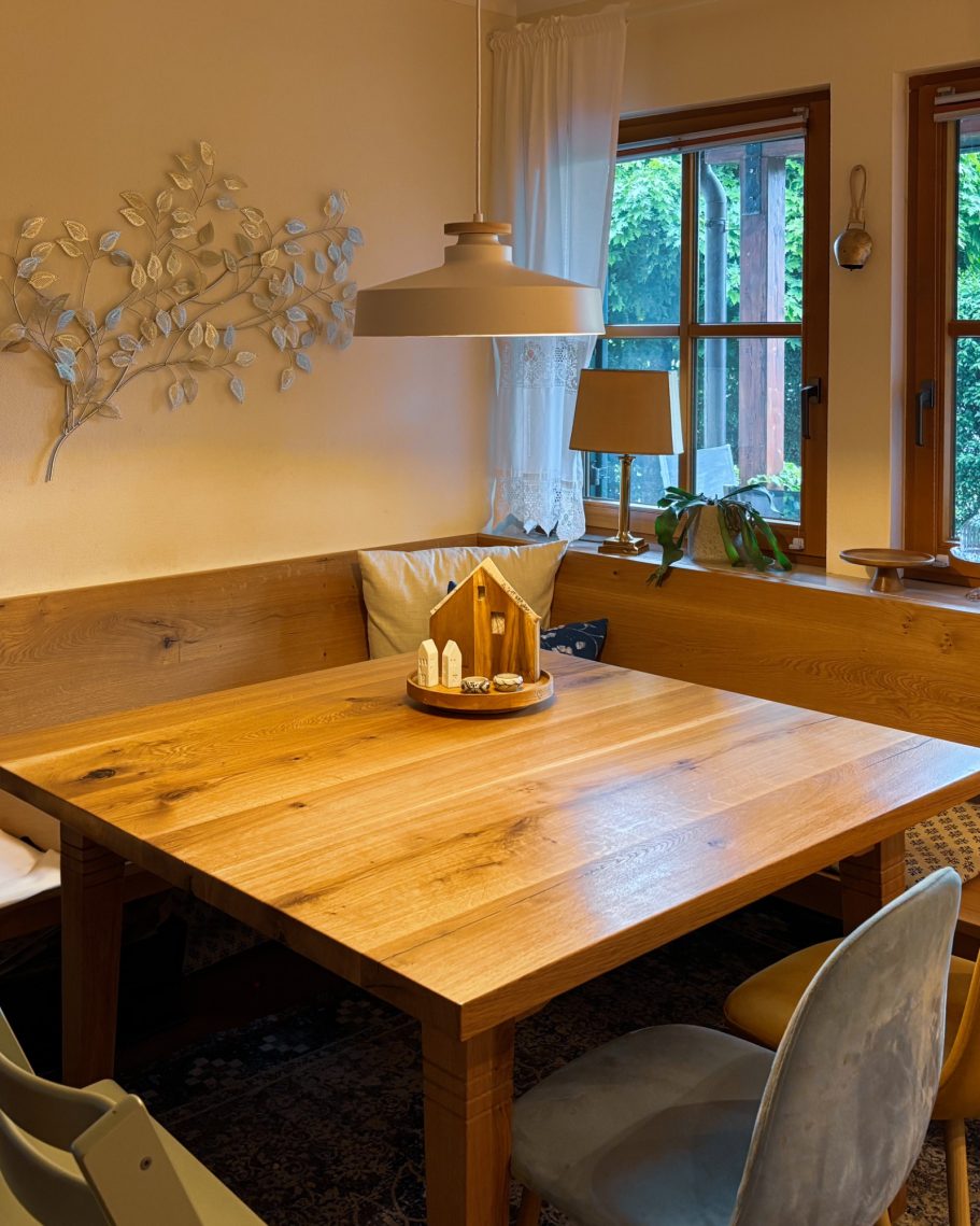 Holztisch mit Sitzgelegenheiten, dekoriert mit einem Holzhaus und Pflanzen im Hintergrund.